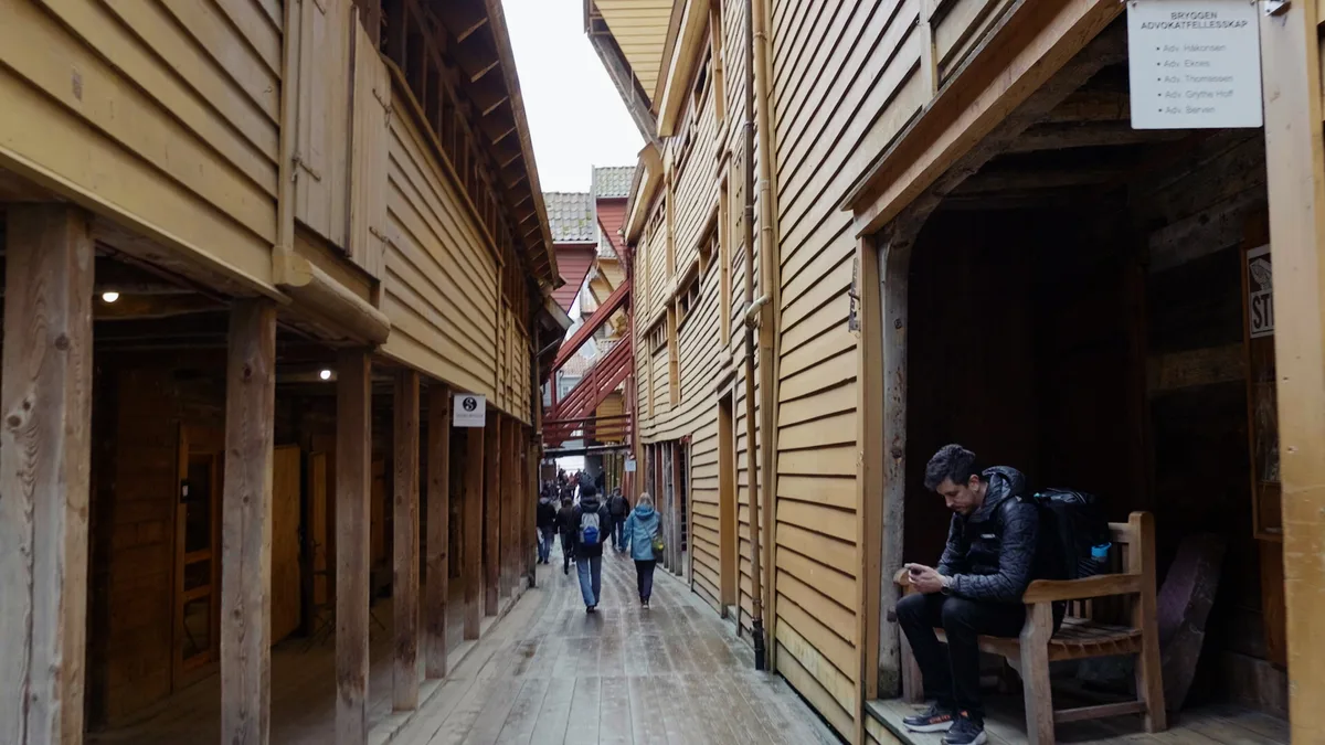 木造の建物に挟まれた、板張りの細い通路。軒下に人が腰かけている